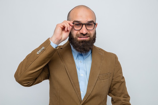 Hombre barbudo en traje marrón con gafas mirando sonriendo alegremente tocando sus gafas