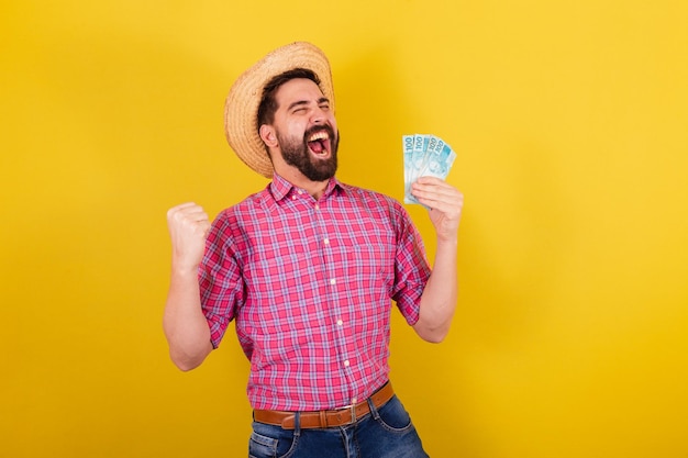 Hombre barbudo vestido con ropa típica para la fiesta Junina Corriendo y celebrando la celebración de la victoria de los billetes de dinero Para la Fiesta de Arraia