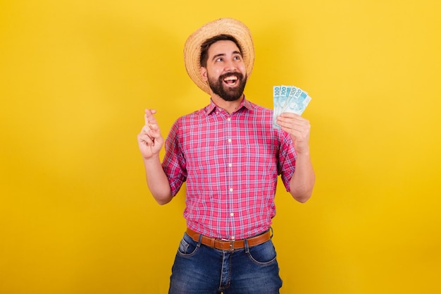 Hombre barbudo vestido con ropa típica para la fiesta Junina cruzando los dedos retorciendo y deseando tener dinero dinero billetes suerte para la fiesta de Arraia