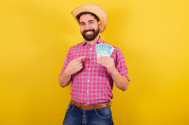 Hombre barbudo vestido con ropa típica para la fiesta Junina hombre señalando billetes de dinero para la fiesta de Arraia