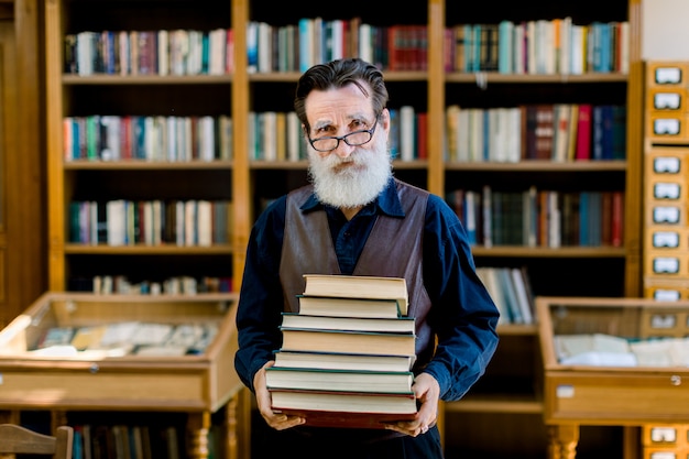 Hombre barbudo viejo inteligente positivo en camisa oscura y chaleco de cuero, trabajador de la biblioteca, profesor, trabajando en la biblioteca, sosteniendo la pila de libros mientras está de pie sobre el fondo de los estantes de libros