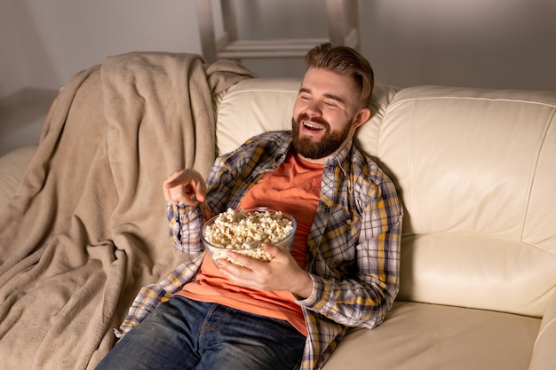 Hombre barbudo viendo películas o juegos deportivos TV comiendo palomitas de maíz en casa por la noche. Cine, campeonato