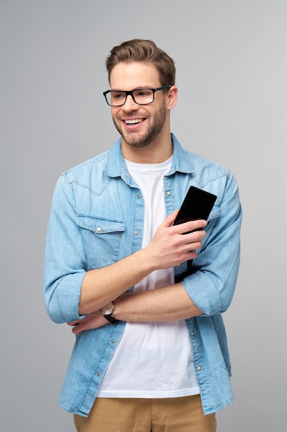 Hombre bastante casual en camisa de jeans azul sosteniendo su teléfono de pie sobre la pared gris del estudio