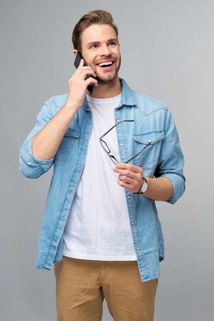 Hombre bastante casual en camisa de jeans azul sosteniendo su teléfono de pie sobre la pared gris del estudio