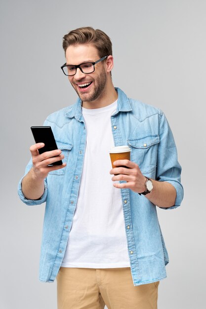 Hombre bastante casual en camisa de jeans azul sosteniendo su teléfono y una taza de café para ir de pie sobre la pared gris del estudio