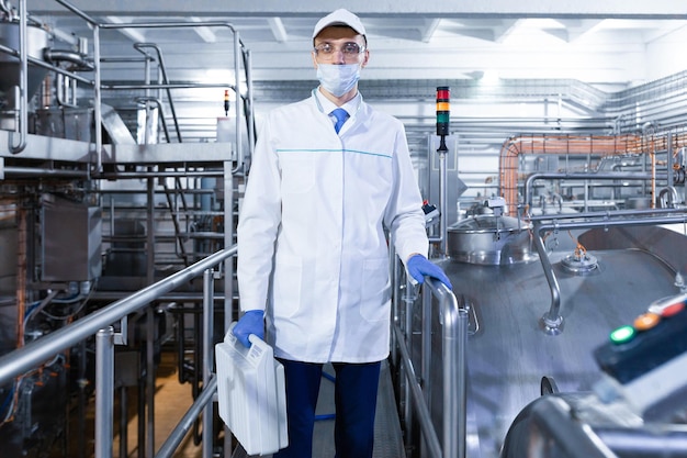 Un hombre con bata blanca, guantes y máscara se para cerca de la línea de producción y sostiene una maleta blanca en la mano en el inspector de la planta de productos lácteos que lleva a cabo el control en la fábrica de queso