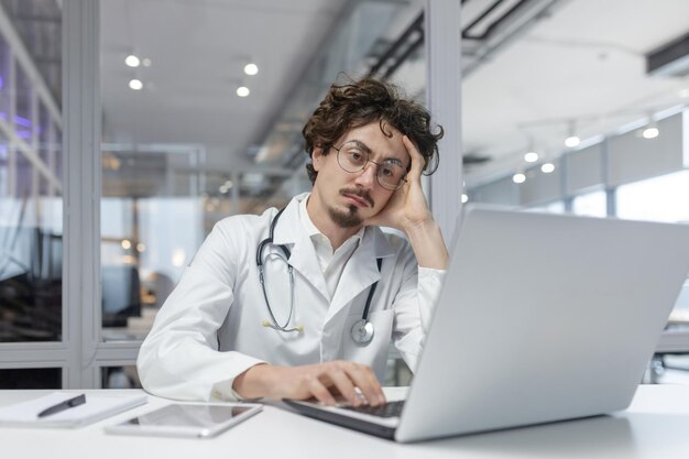 Un hombre con una bata médica blanca con un estetoscopio se sienta frente a una computadora portátil en un médico