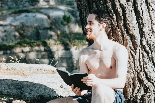 Un hombre en baúles sin camisa leyendo un libro contra un árbol durante un día de verano, relajarse y relajarse conceptos, buena vida, gente joven, mirando lejos del espacio de copia de la cámara