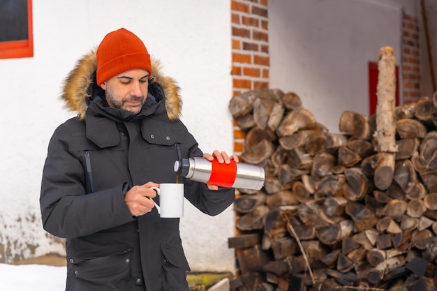 Hombre bebiendo café de un termo caliente en invierno en la nieve junto a la madera de la chimenea