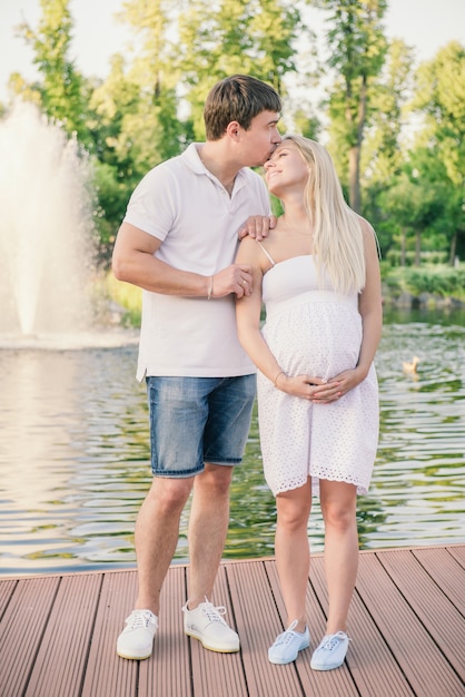 Hombre besando a feliz mujer embarazada en el muelle cerca del agua por la noche. La nueva familia se apoya mutuamente.