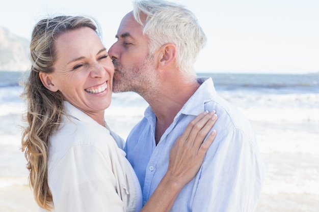 Hombre besando a su compañero sonriente en la mejilla en la playa