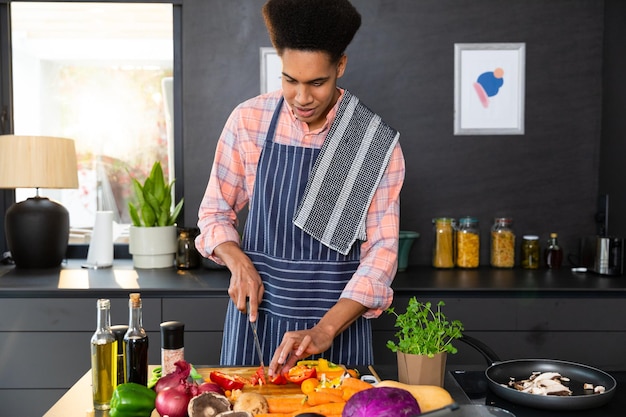 Hombre bi-racial feliz con delantal preparando comida, cortando verduras en una cocina soleada. Comida, cocina, estilo de vida saludable y vida doméstica, inalterados.