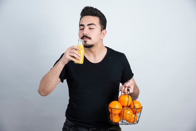 Hombre con bigote que sostiene una botella de jugo de vidrio con una canasta metálica llena de frutas naranjas.