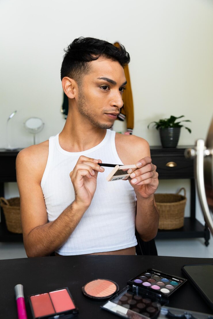 Foto hombre biracial transgénero mirándose en el espejo y poniéndose maquillaje, aplicándose sombra de ojos. género, cuidado personal, belleza, estilo de vida y vida doméstica, inalterados.