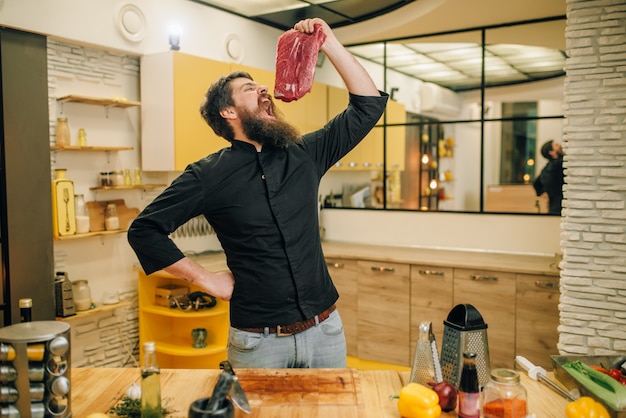 El hombre con la boca abierta sostiene un trozo de carne cruda fresca. Chef cocinando solomillo con verduras, especias y hierbas.