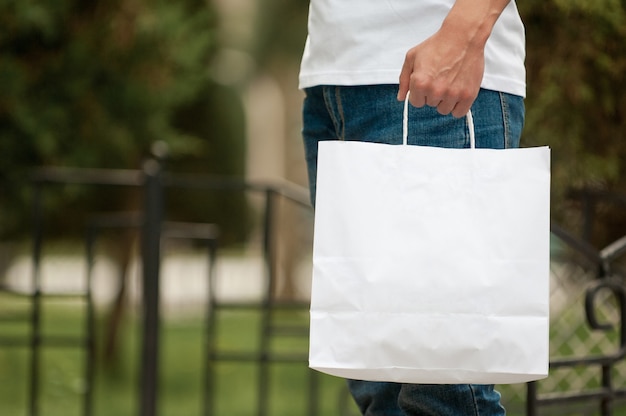 Hombre con una bolsa de papel blanco en las manos sobre un fondo de parque. Mensajero con una bolsa en el fondo de un parque verde