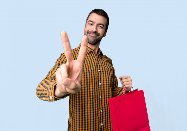 Foto el hombre con los bolsos de compras que sonríe y que muestra la victoria firma en fondo azul aislado