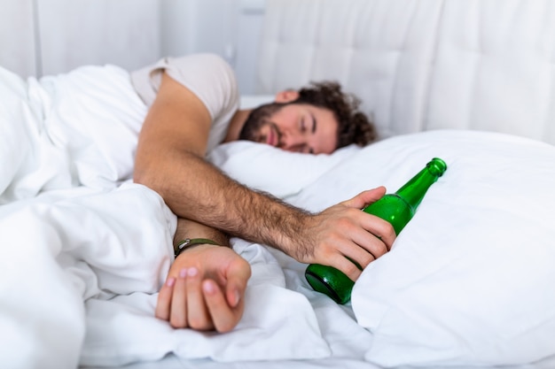 Hombre borracho en la cama y triste lugar y una botella de alcohol en la mano. Hombre joven que miente en la cama borracho mortal que sostiene la botella casi vacía de alcohol.