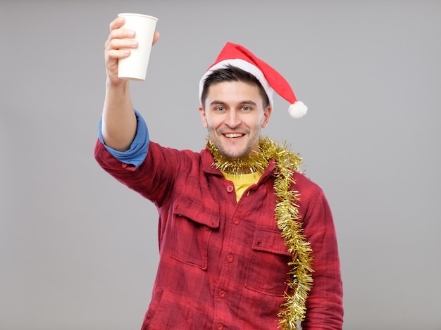 Hombre borracho joven divertido que lleva el sombrero de Papá Noel que sostiene una taza de papel