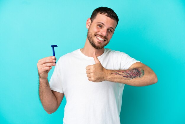 Hombre brasileño afeitándose la barba aislado sobre fondo azul dando un pulgar hacia arriba gesto