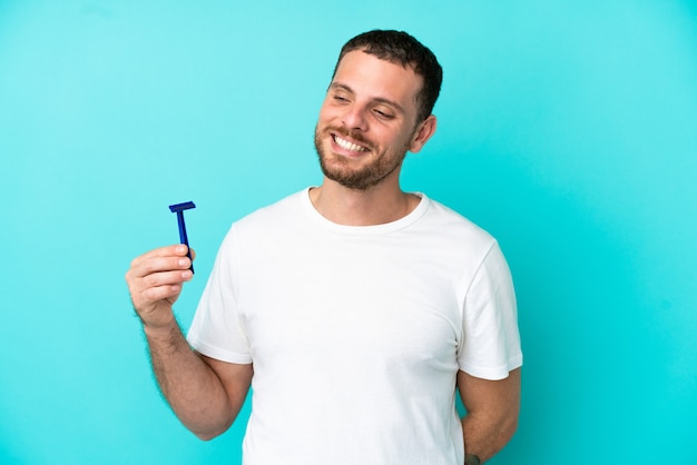 Hombre brasileño afeitándose la barba aislado sobre fondo azul mirando hacia el lado y sonriendo