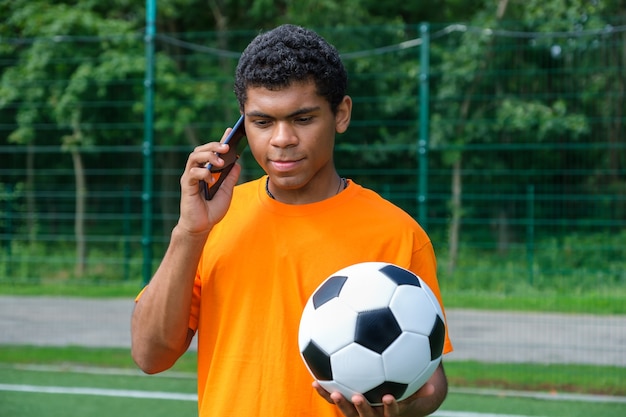 Hombre brasileño que sostiene un balón de fútbol y usa un teléfono inteligente en la cancha deportiva