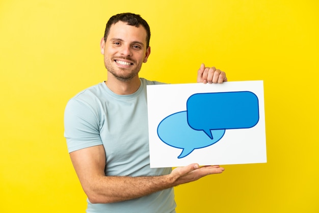 Hombre brasileño sobre fondo púrpura aislado sosteniendo un cartel con el icono de la burbuja del discurso con expresión feliz