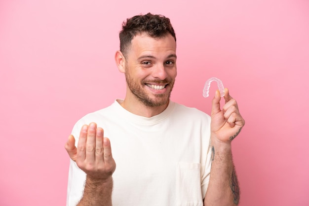 Hombre brasileño sosteniendo aparatos ortopédicos invisibles invitando a venir con la mano Feliz de que hayas venido