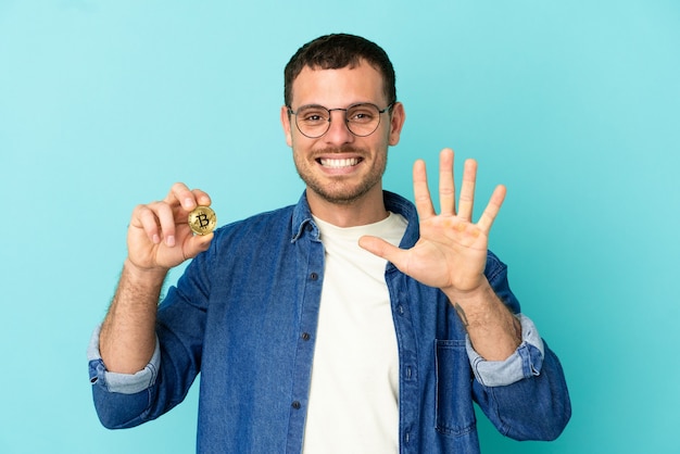Foto hombre brasileño sosteniendo un bitcoin sobre fondo azul aislado contando cinco con los dedos