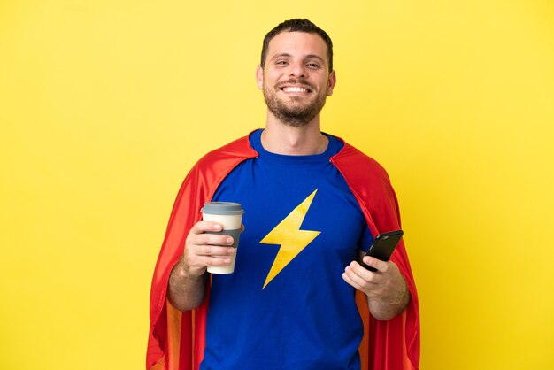 Hombre brasileño de superhéroe aislado sobre fondo amarillo con café para llevar y un móvil