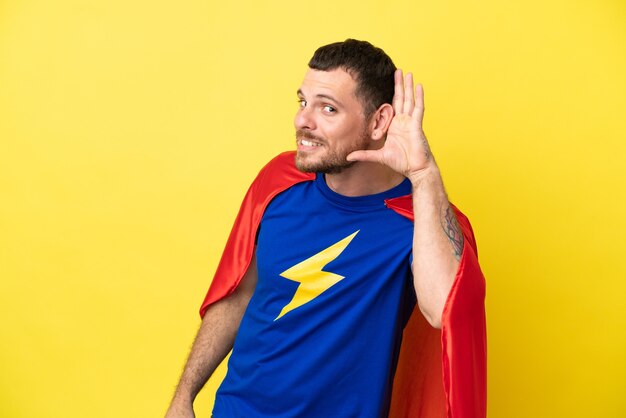 Hombre brasileño de superhéroe aislado sobre fondo amarillo escuchando algo poniendo la mano en la oreja