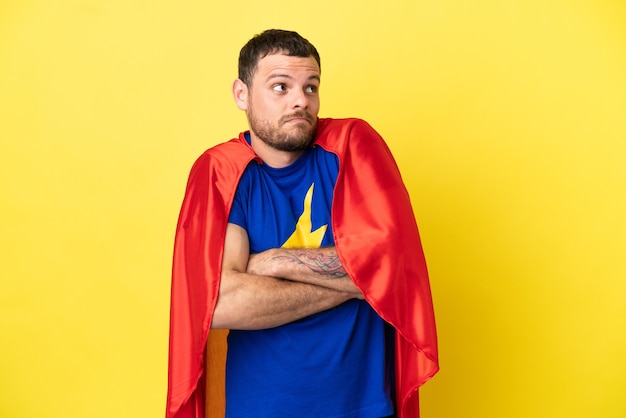 Hombre brasileño de superhéroe aislado sobre fondo amarillo haciendo gesto de dudas mientras levanta los hombros
