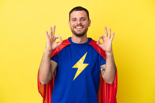 Hombre brasileño de superhéroe aislado sobre fondo amarillo mostrando un signo de ok con los dedos