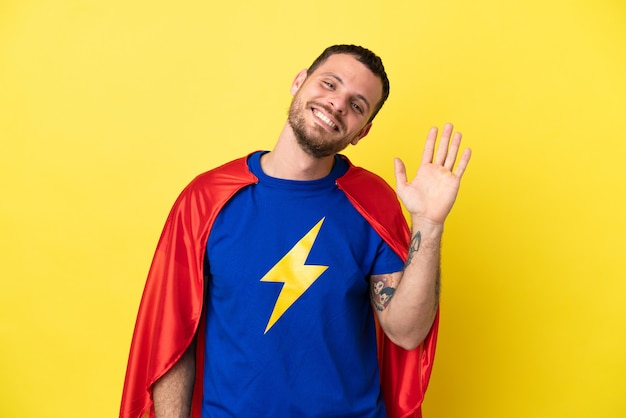 Hombre brasileño de superhéroe aislado sobre fondo amarillo saludando con la mano con expresión feliz