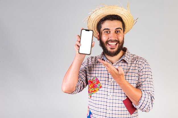 Hombre brasileño vestido con ropa de festa junina Arraial Fiesta de San Juan con smartphone y pantalla blanca