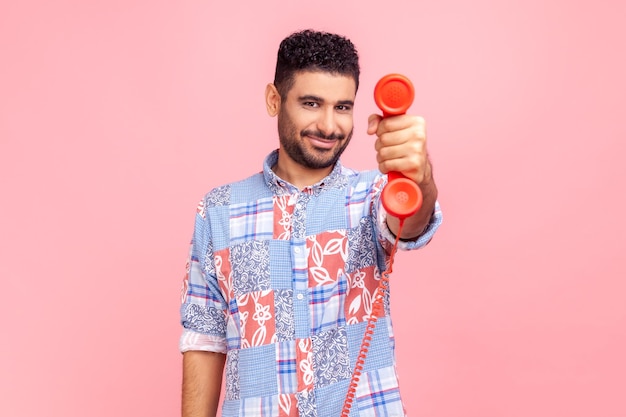 Hombre brunet con barba con camisa azul de estilo informal sosteniendo y mostrando un teléfono fijo, mirando la cámara con una sonrisa. Disparo de estudio interior aislado sobre fondo rosa.
