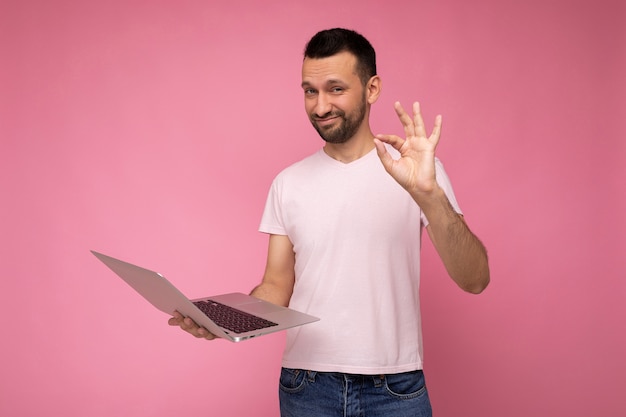 Hombre brunet divertido hermoso que sostiene la computadora portátil y que muestra el gesto bien mirando a la cámara en la camiseta