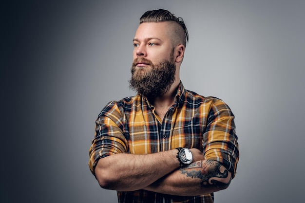 Hombre brutal barbudo con brazos cruzados tatuados sobre fondo gris.