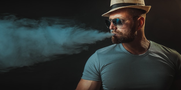 Hombre brutal barbudo con sombrero y gafas de sol fumando un cigarrillo de vapor como alternativa al tabaco en negro