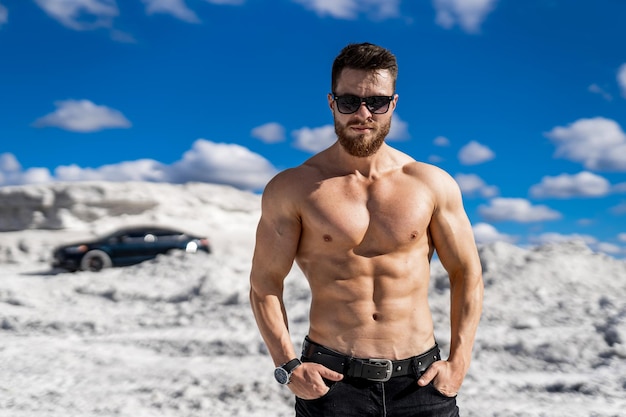 Hombre brutal con cuerpo perfecto y abdominales fuertes usa gafas de sol y mira a la cámara Cantera blanca y fondo de auto negro de lujo