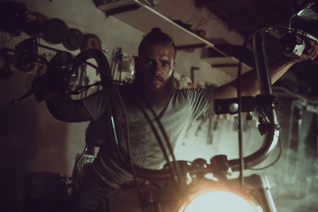 Hombre brutal guapo con barba sentado en una motocicleta en su garaje, limpiándose las manos y mirando a un lado