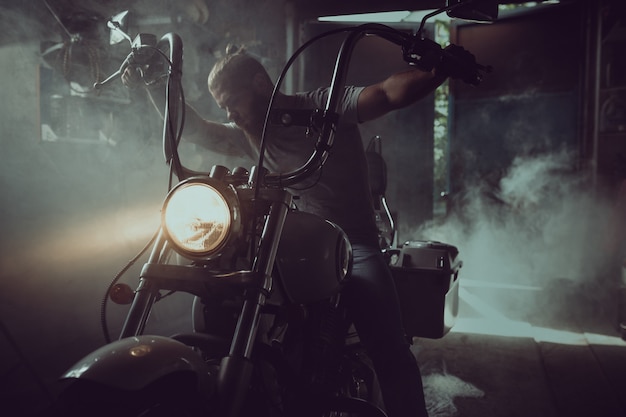 Hombre brutal guapo con barba sentado en una motocicleta en su garaje