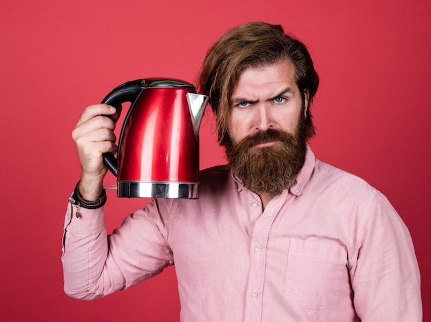 Hombre brutal serio con bigote y barba dispositivo doméstico eléctrico hombre barbudo usa hervidor eléctrico rojo concepto de limpieza masculino necesita un poco de agua caliente hombre que va a beber té o café