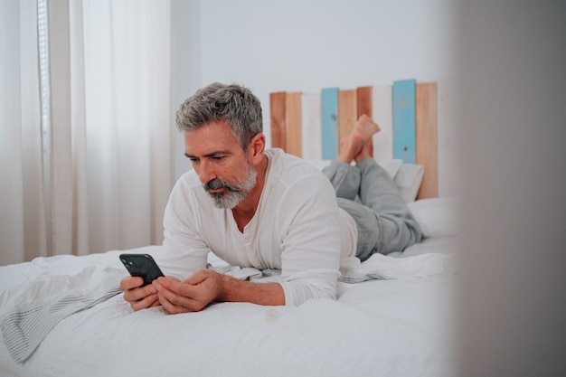 Hombre con cabello gris de unos 50 años acostado en su cama mirando el teléfono al levantarse