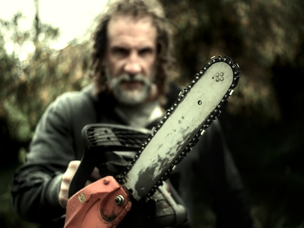 Foto hombre con cabello largo y barba en el fondo borroso sosteniendo una motosierra enfocada en primer plano