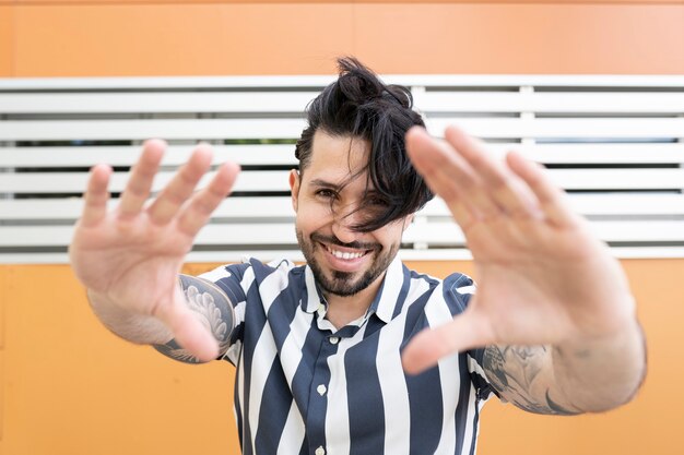 Hombre con cabello largo y tatuajes, con bonita sonrisa, pone sus manos en gesto de foto