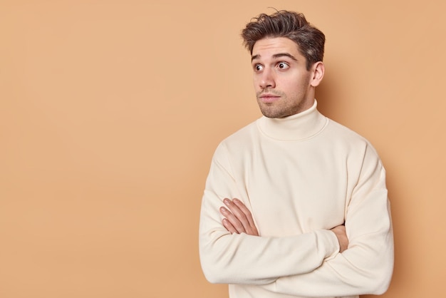 hombre con cabello oscuro mantiene los brazos cruzados mira sorprendentemente lejos reacciona en una escena impactante usa un jersey casual aislado en una pared beige con espacio de copia en blanco.