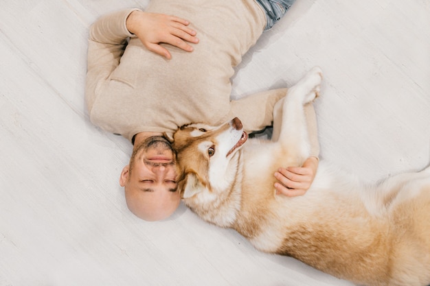 Hombre calvo adulto con cachorro husky durmiendo en el piso. Propietario con mascota juntos en casa. Emociones amables y conmovedoras. Precioso perro descansando con macho joven. Chico con amado animal doméstico abrazándose unos a otros.