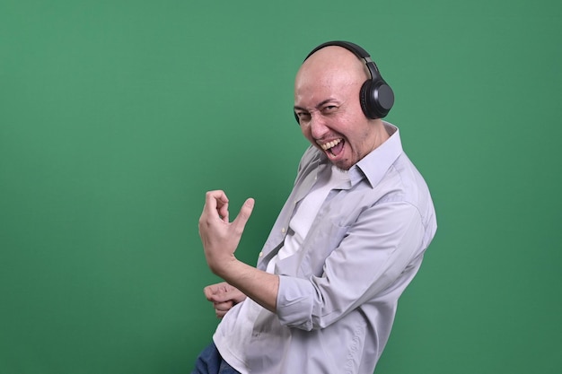 Hombre calvo asiático escuchando música con auriculares mientras hace un gesto de guitarra sobre un fondo verde