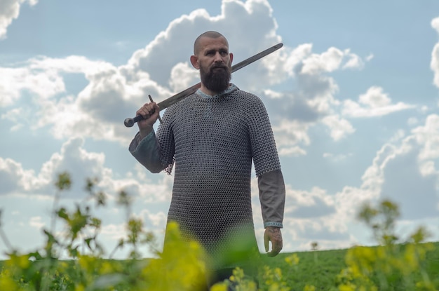 Un hombre calvo con barba y una cota de malla metálica sobre una camisa de lino se encuentra en medio del campo sosteniendo una espada